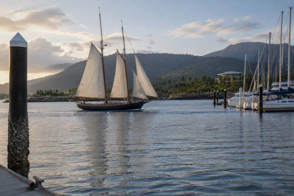 A timber sailing schooner under way on blue tropical water with a forested island in the distance