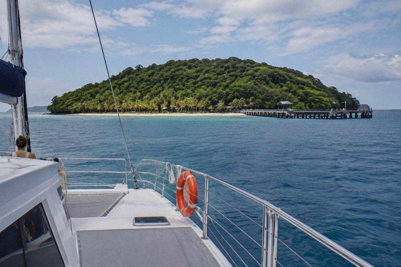 A large catamaran cruising across calm blue tropical water toward a green tree covered coral cay