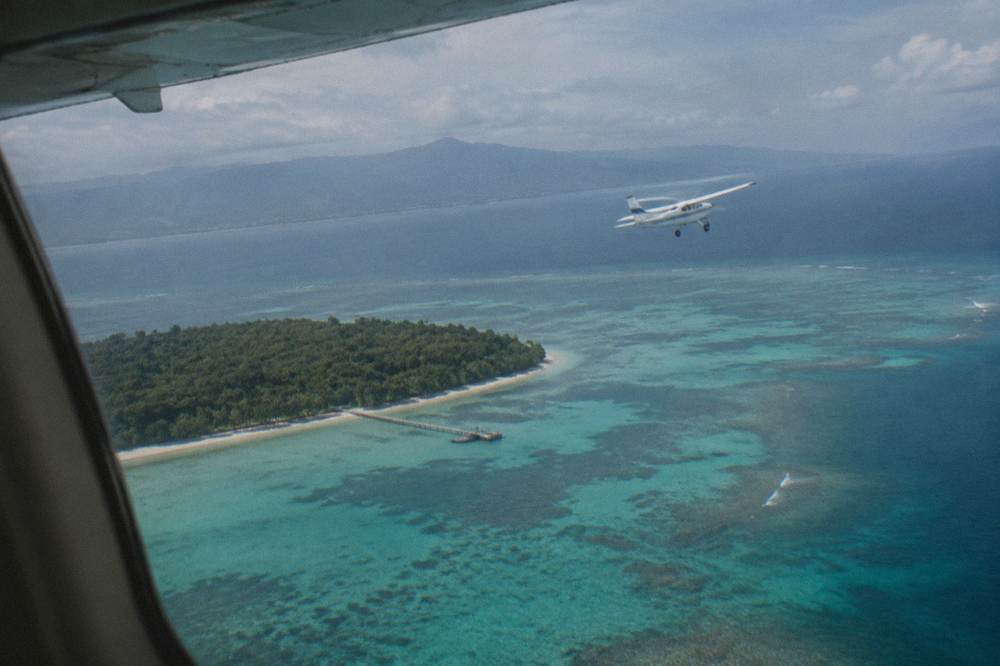 A high-wing light aircraft flying over reef and turquoise water near an island