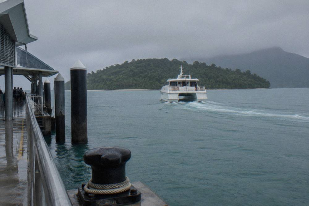 A white passenger catamaran crossing calm turquoise water toward a green coral island