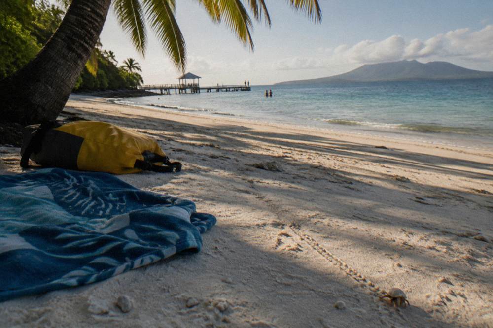 A beach towel and dry bag on white sand under palm shade near calm water