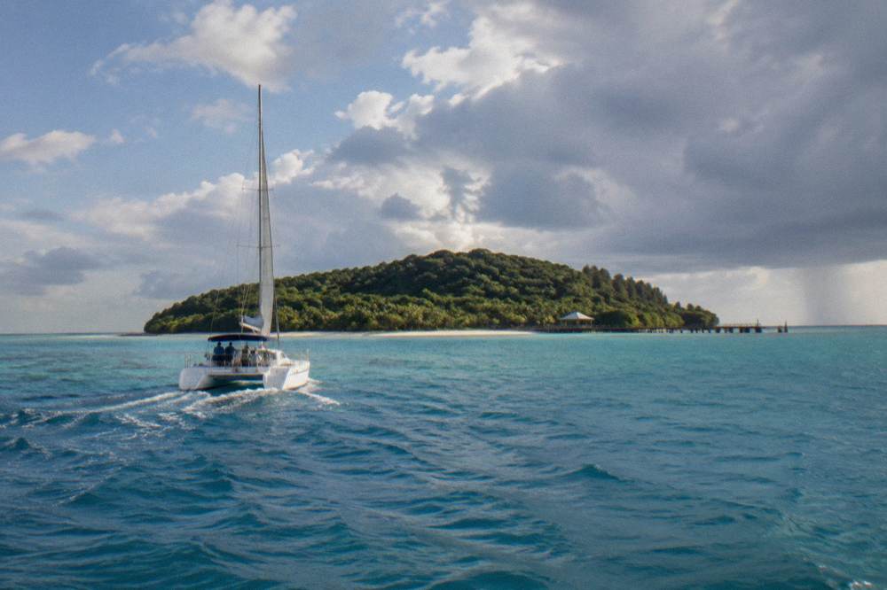 A catamaran cruising towards a green tropical island under a mix of sun and cloud