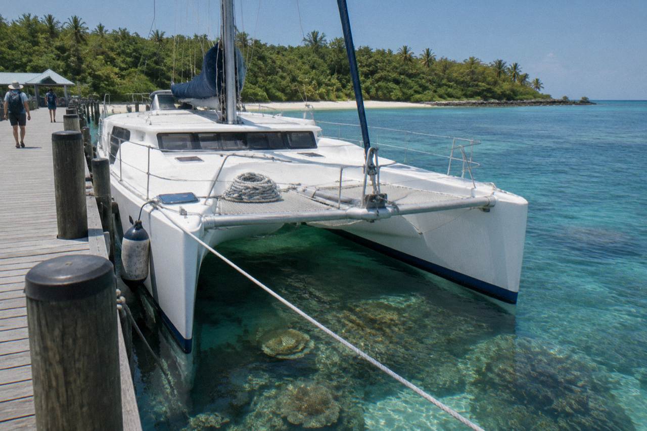 A large catamaran moored at a tropical island jetty beside clear shallow reef water