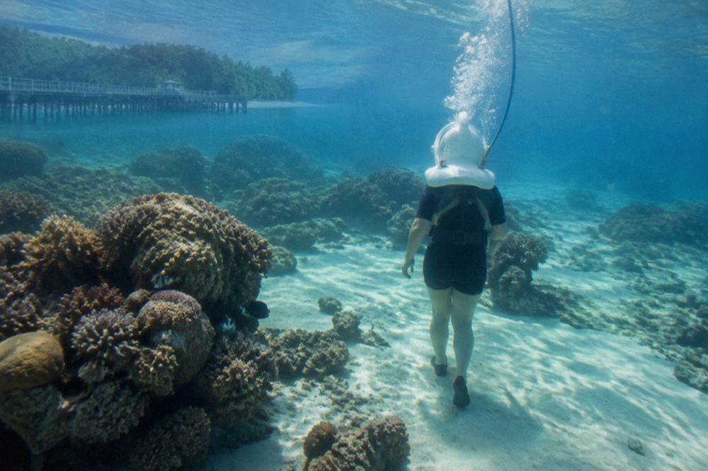 A walker in a diving helmet standing on a sandy seabed among coral