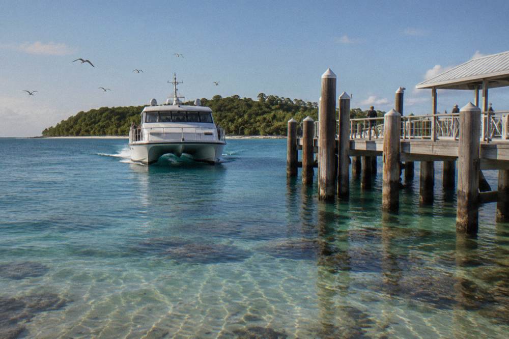 A white catamaran ferry approaching a timber jetty over clear shallow reef water