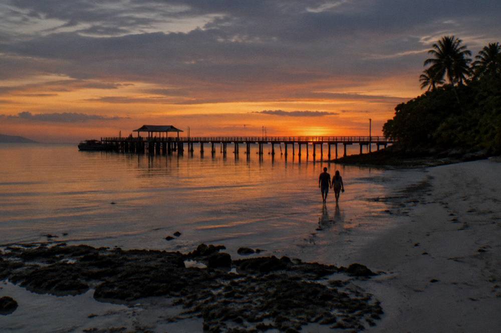 A calm sandy beach at dusk with a timber jetty silhouette and darkening sky over reef water