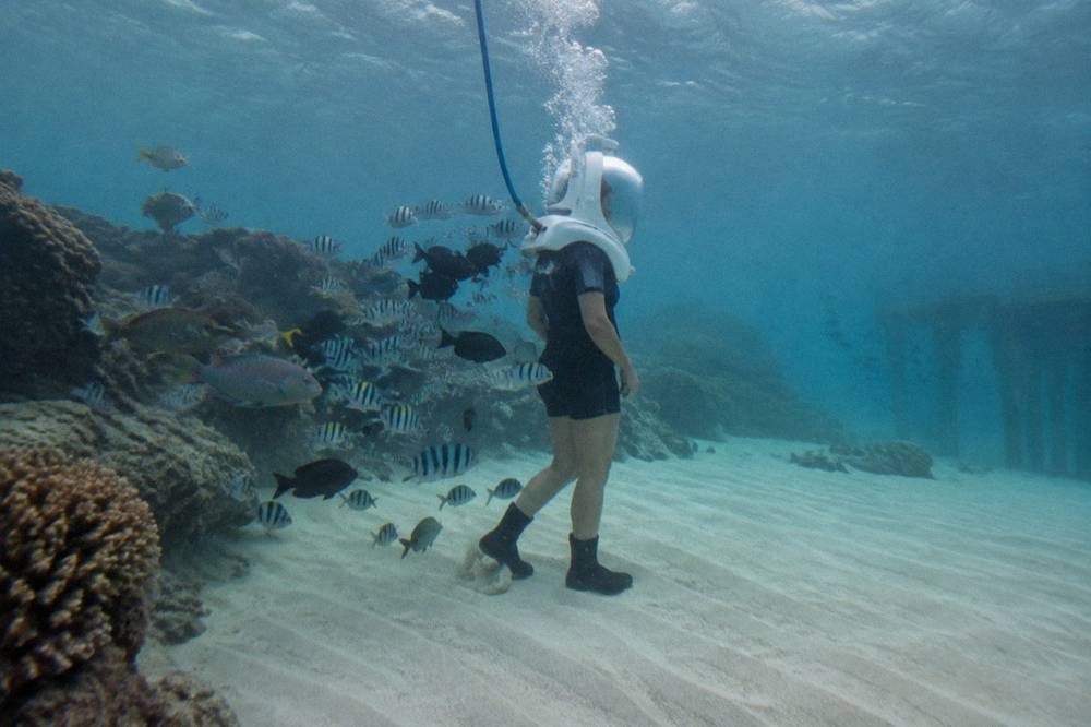 A walker in a sealed diving helmet standing on a sandy reef floor surrounded by tropical fish