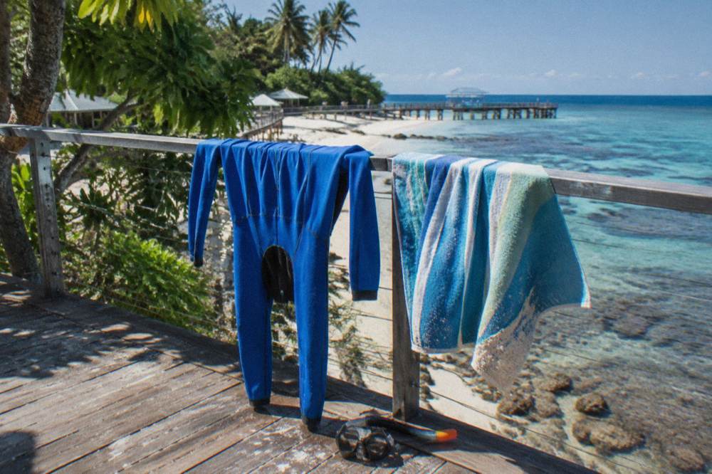 A full body lycra stinger suit drying on a railing beside a beach towel and mask