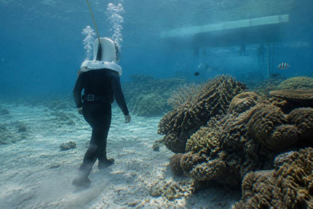A diver in a helmet walking on a sandy seabed beside coral in clear water