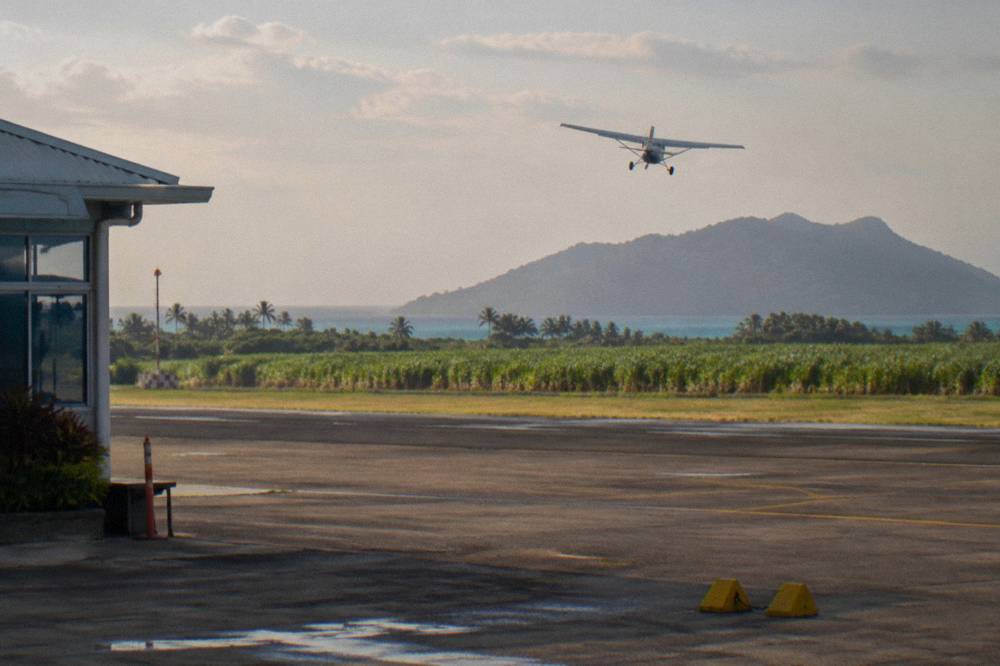 A high-wing light aircraft flying over turquoise reef water and a small green island
